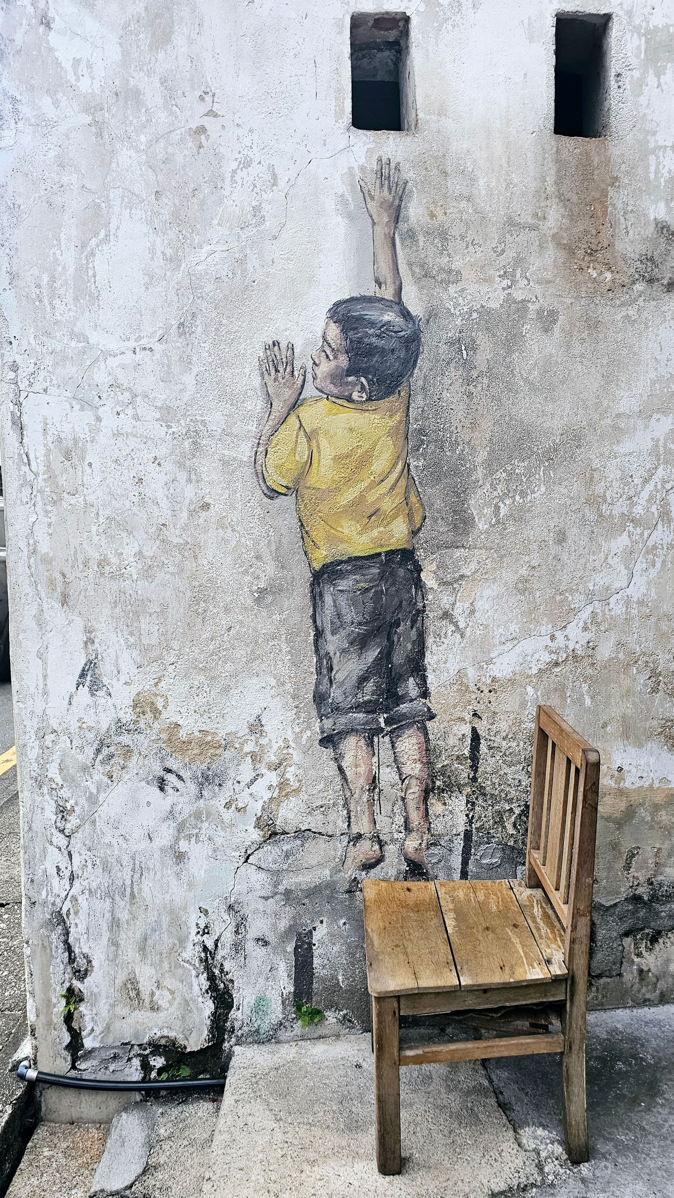 Reaching Up mural by Ernest Zacharevic on Cannon Street, a painted boy standing on a real wooden chair to reach a window