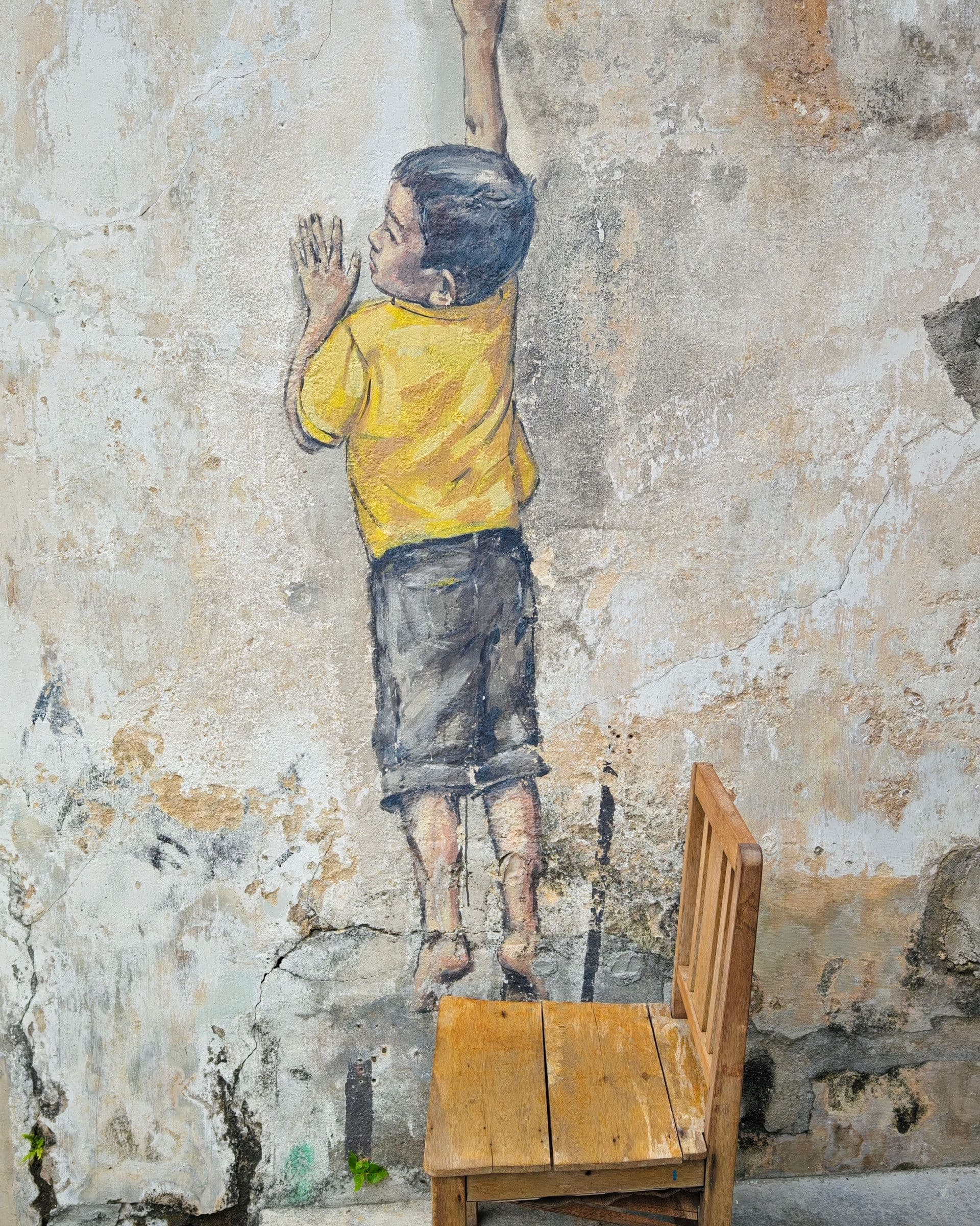 Reaching Up mural by Ernest Zacharevic on Cannon Street, a painted boy standing on a real wooden chair reaching toward a window